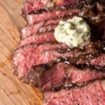 Sliced medium-rare reverse sear ribeye on a wooden cutting board, topped with a dollop of ultimate cowboy butter and sprinkled with coarse salt.