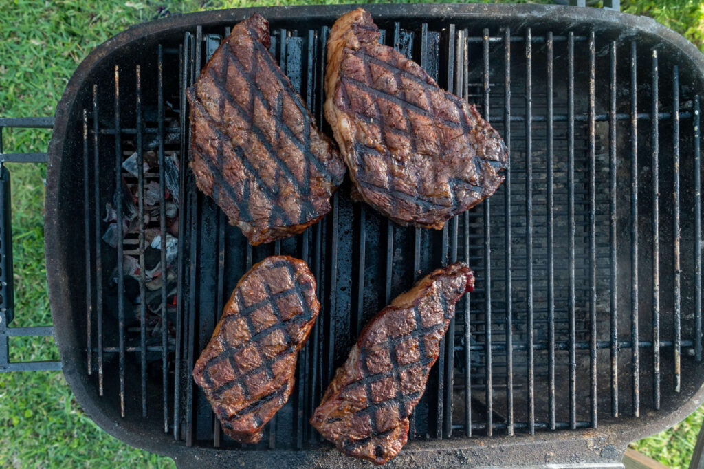 Four grilled steaks with char marks sizzle on an outdoor barbecue grill over hot coals, perfect for pairing with the ultimate Cowboy Butter recipe. Fresh green grass is visible in the background.