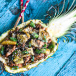 A pineapple bowl of teriyaki chicken and pineapple pieces topped with sesame seeds and scallions.