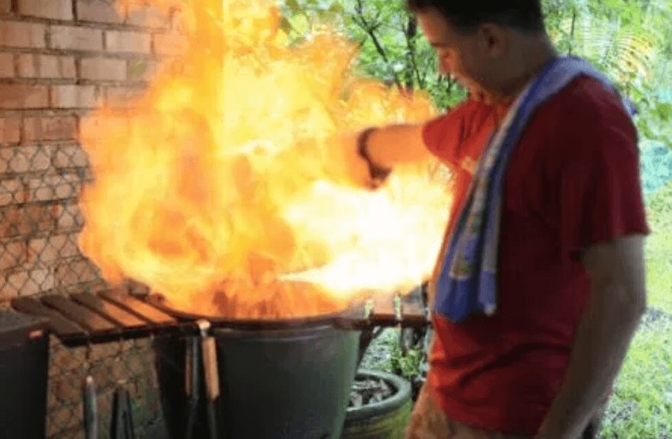 A person in a red shirt stands near a grill with a large burst of flames, possibly demonstrating how to control temp on the Big Green Egg, holding a cooking tool with a towel over their shoulder, next to a brick wall and some greenery.