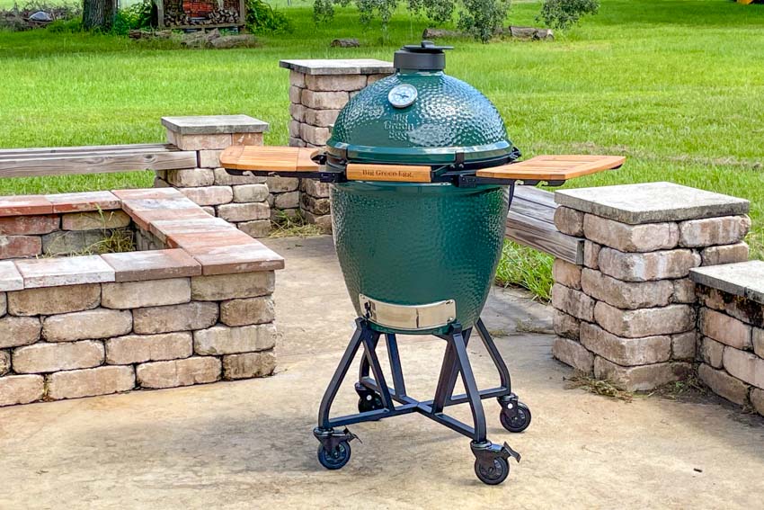 A green ceramic outdoor grill and smoker, known as a Big Green Egg, stands on a wheeled metal cart with wooden side shelves in a stone patio area surrounded by a green lawn—perfect for learning how to control temp on the Big Green Egg.