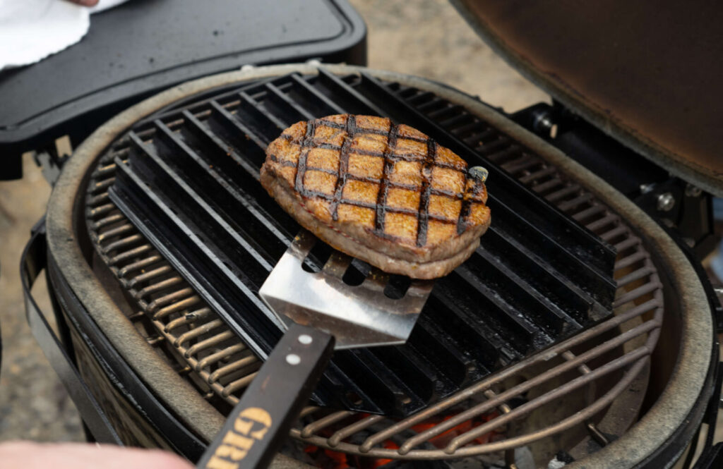 A person grills a SCA Steak on an outdoor barbecue, using a spatula to lift the meat, which shows distinct grill marks. A black tray with a white towel is beside the grill.
