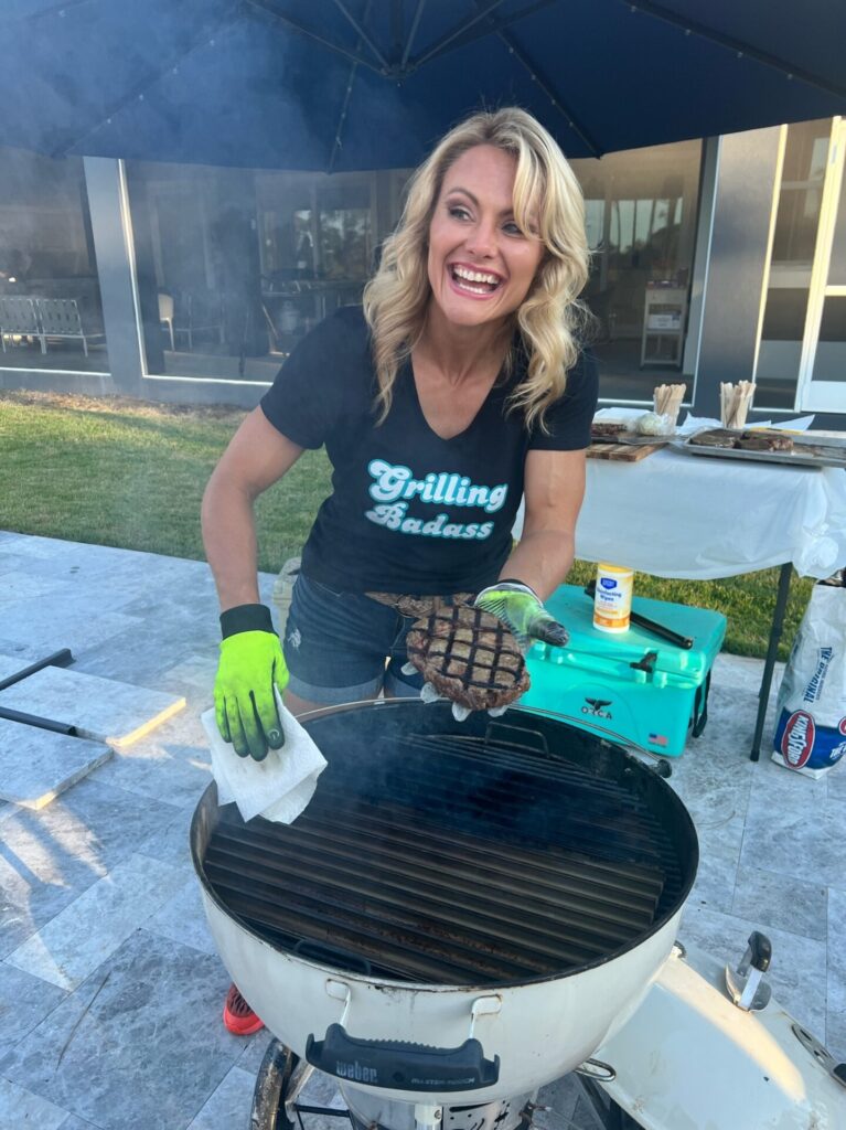 A smiling woman wearing a Grilling Badass shirt and green gloves proudly holds an SCA Steak above a barbecue grill outdoors, with picnic tables, food, and a cooler in the background.