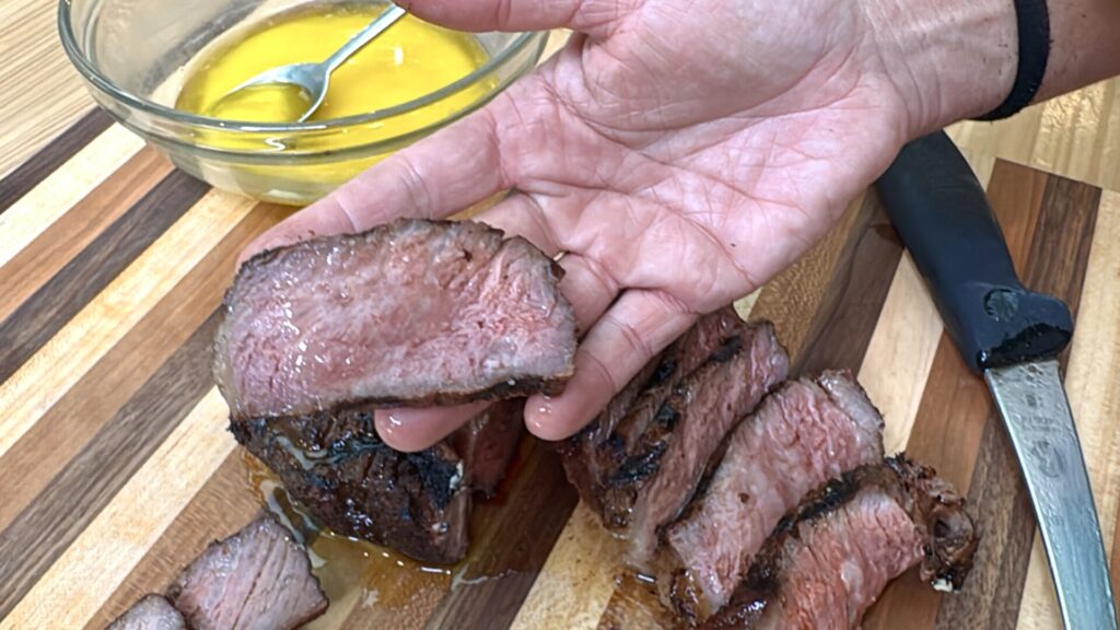A hand holds a juicy, medium-cooked SCA Steak slice over a wooden cutting board, with more sliced pieces nearby and a bowl of melted butter with a spoon in the background.