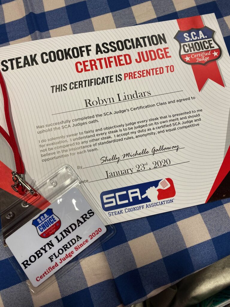 A certificate and name badge for Robyn Lindars, showing they are a certified SCA Steak judge for the Steak Cookoff Association, displayed on a blue and white checkered tablecloth. The certificate is dated January 23, 2020.