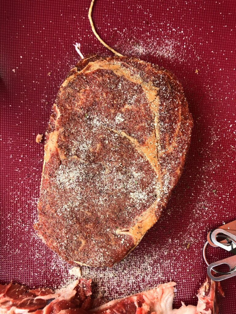A raw, seasoned SCA Steak rests on a red cutting board, covered with a generous layer of salt and pepper. A few pieces of trimmed meat and a metal utensil are visible near the bottom right corner.