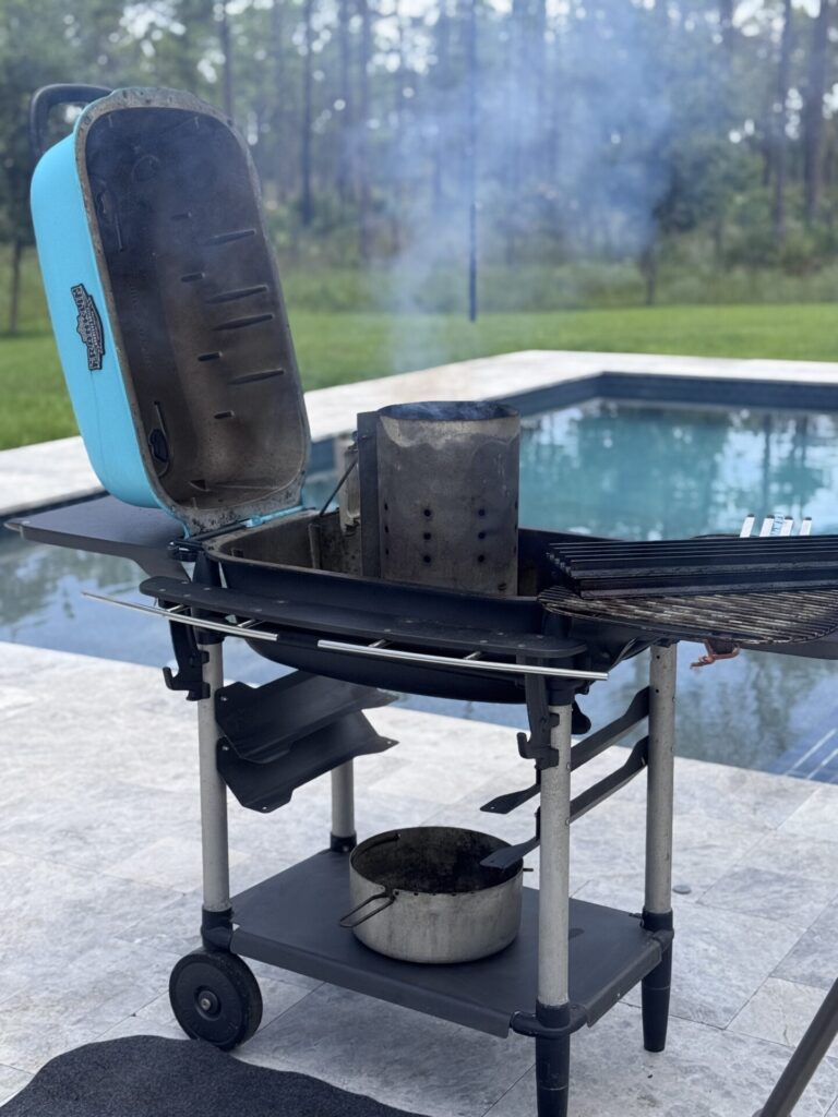 A charcoal grill with its lid open sits by a pool, smoke rising from a chimney starter inside—perfect for your next SCA Steak cook-off. A pot rests on the shelf below, framed by trees in the background.