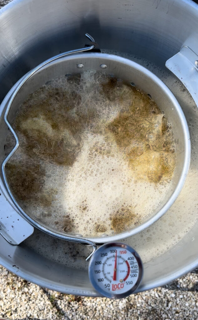A large metal pot filled with bubbling hot oil is frying food outdoors on a gravel surface. A thermometer tracks the heat as a round basket holds what could be a Holiday Obsession favorite&mdash;perhaps even a Smoke-Fried Turkey sizzling to perfection.