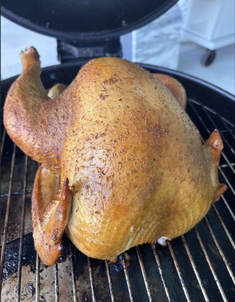 A whole, golden-brown chicken is standing upright on a grill inside a barbecue smoker, being cooked outdoors&mdash;a true Holiday Obsession, with skin that's crispy, evenly roasted, and perfectly seasoned.
