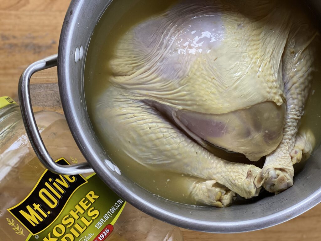 A whole raw chicken sits in a pot filled with brine, possibly infused with pickle juice from the Mt. Olive Kosher Dills jar nearby. This setup could be perfect for preparing a tender holiday turkey or even a flavorful smoke-fried turkey.