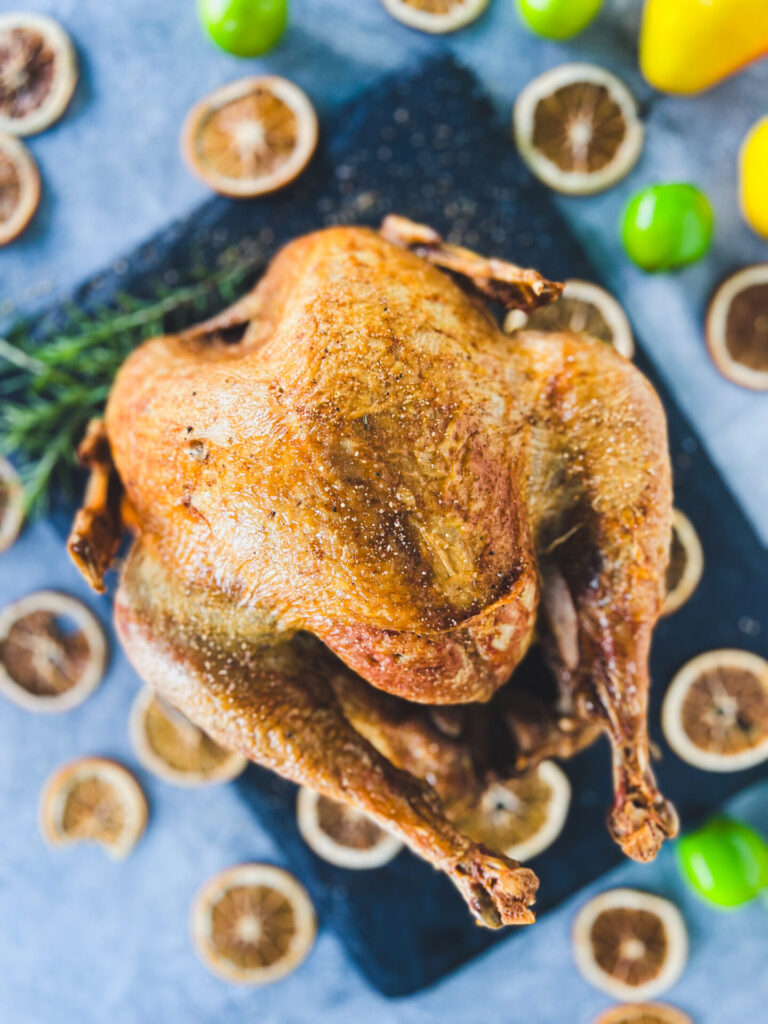 A whole smoke-fried turkey sits on a dark tray, surrounded by slices of dried lemon and fresh green limes on a blue surface.