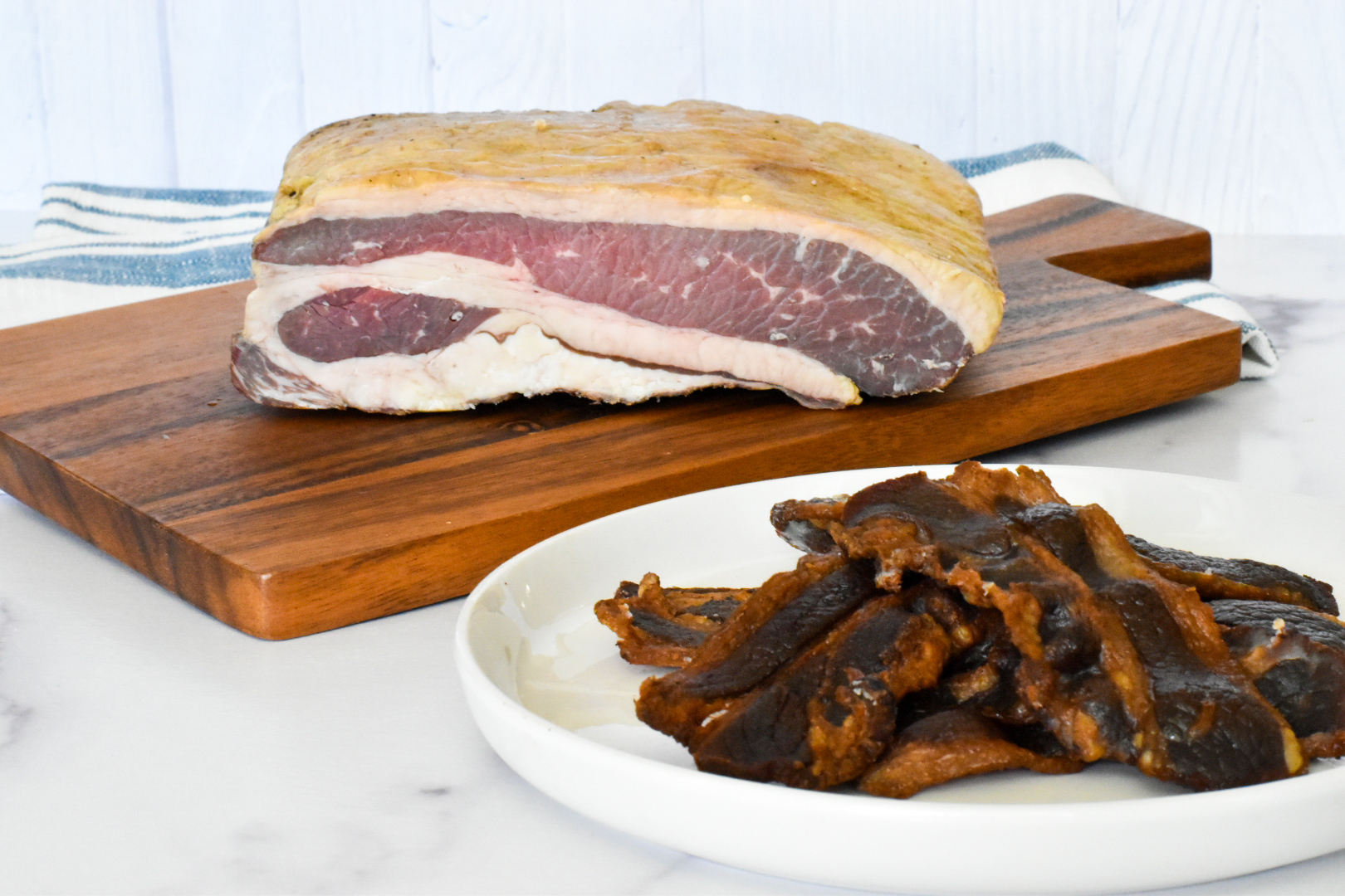 A thick slab of Naturally Cured Beef Bacon sits on a wooden cutting board, while several cooked bacon strips are arranged on a white plate in the foreground. A striped towel is in the background.