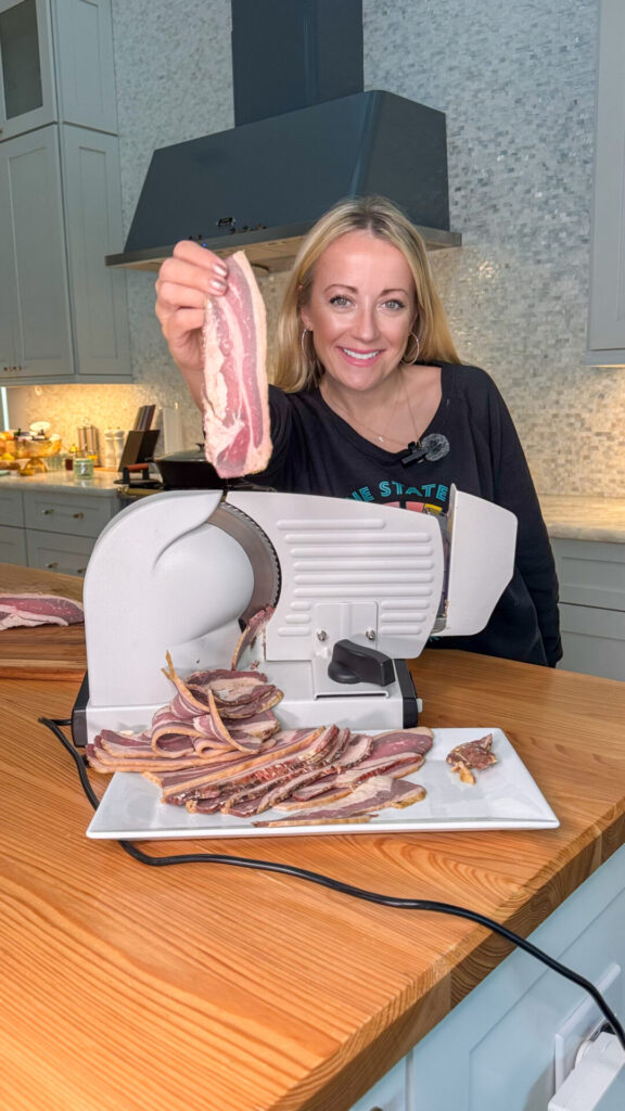 A smiling woman in a kitchen holds up a slice of naturally cured beef bacon next to a meat slicer, with several bacon slices arranged on a white plate on the wooden countertop in front of her.